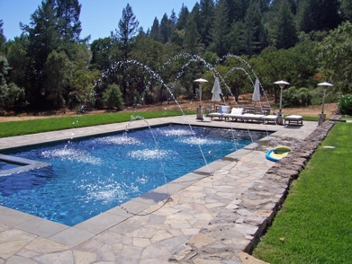 A swimming pool with arc fountains of water over it and a stone 